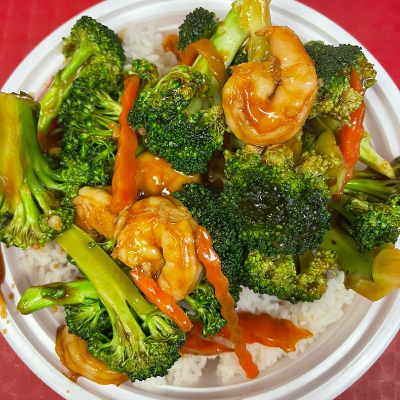 Shrimp and broccoli stir-fry with carrots and a savory sauce, served over white rice in a takeout container.